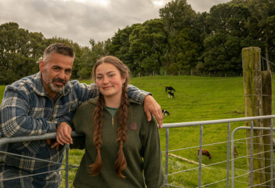 An older man and a young woman lean against a steel gate on a pasture with cows in the background.