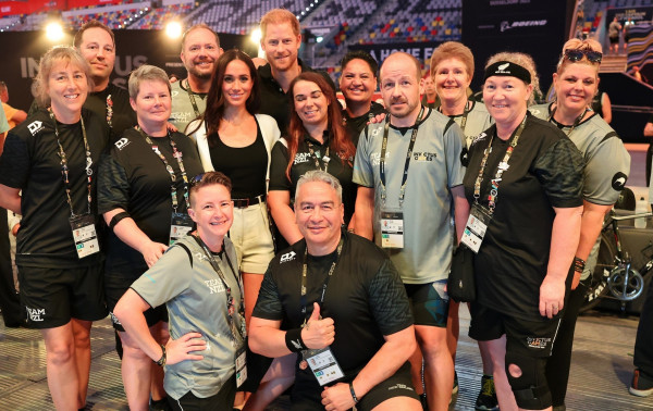 Prince Harry and Meghan Markle standing among members from the New Zealand Team in a sports stadium. 