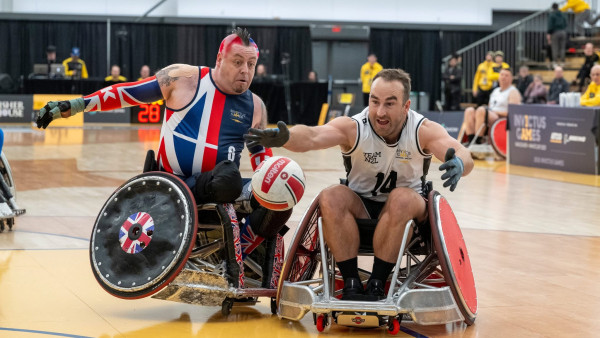Two men in sport wheelchairs competing for a ball in a basketball stadium.