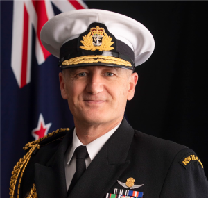 A headshot of a man in ceremonial navy uniform with a New Zealand flag in the background