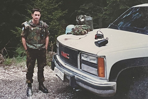 A man in Army uniform standing by a truck on a gravel path.