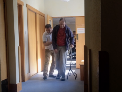 Richard walking with a crutch and the support of a medical professional