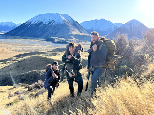 Three men standing on a hill in the mountains carrying deer carcasses over their shoulders