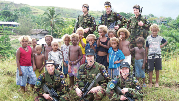 Six NZ Army soldiers smiling in uniform. They are surrounded by 16 young children on a grassy hill